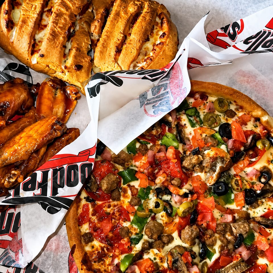 Loaded meat-lover’s pizza, cheesy breadsticks, and saucy chicken wings arranged on branded restaurant paper.